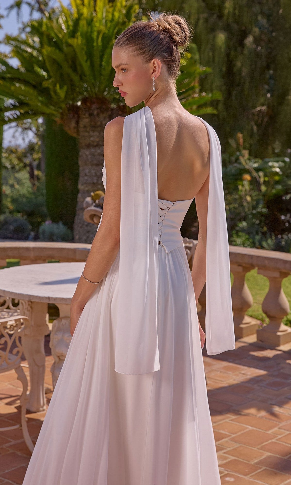 A woman in the Amoris T741W Long Prom Dress with a strapless satin bodice and off-white chiffon A-line skirt stands on a terrace with stone railings, surrounded by lush greenery.